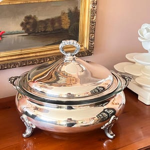 May include: A silver-plated serving dish with a glass insert and a domed lid. The dish has ornate handles and rests on decorative feet. The lid features a decorative handle. The dish is displayed on a wooden surface.