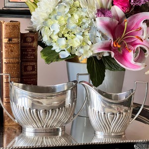 May include: A silver creamer and sugar bowl set with a fluted design, displayed on a silver tray. The set is next to a bouquet of flowers with pink lilies and white hydrangeas. Antique books are in the background.