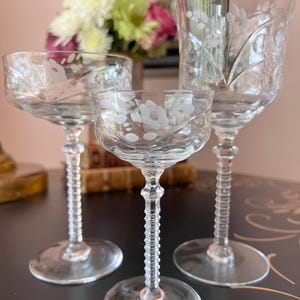 May include: Three clear crystal wine glasses with etched floral designs. The glasses vary in size and stem detail, with a small coupe glass in the center. The stems have a ribbed design. The glasses are on a dark surface.