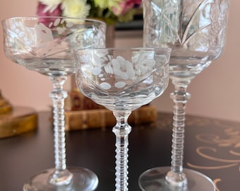 May include: Three clear crystal wine glasses with etched floral designs. The glasses vary in size and stem detail, with a small coupe glass in the center. The stems have a ribbed design. The glasses are on a dark surface.