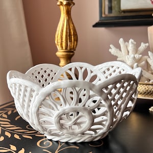 May include: A white ceramic basket with an openwork design. The basket has a scalloped rim and decorative circular and lattice patterns. The basket is sitting on a black surface with gold leaf accents.