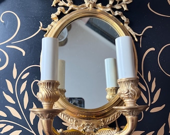 May include: A gold-colored wall sconce with an oval mirror and two faux candle lights. The sconce has ornate floral and scroll detailing. The background is black with a gold leaf pattern.