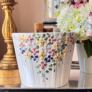 May include: White ceramic planter with embossed floral design in yellow, pink, red, and blue. The planter has vertical ribbed detailing and a slightly flared shape. A decorative piece for indoor plants.