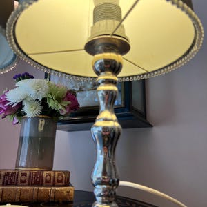 K&ouml;nnte beinhalten: Eine silberne Tischlampe mit beigem Lampenschirm und Perlenbesatz. Die Lampe steht auf einem Tisch mit einer Blumenvase und einem Stapel B&uuml;cher. Die Lampe ist eingeschaltet und wirft ein warmes Licht.