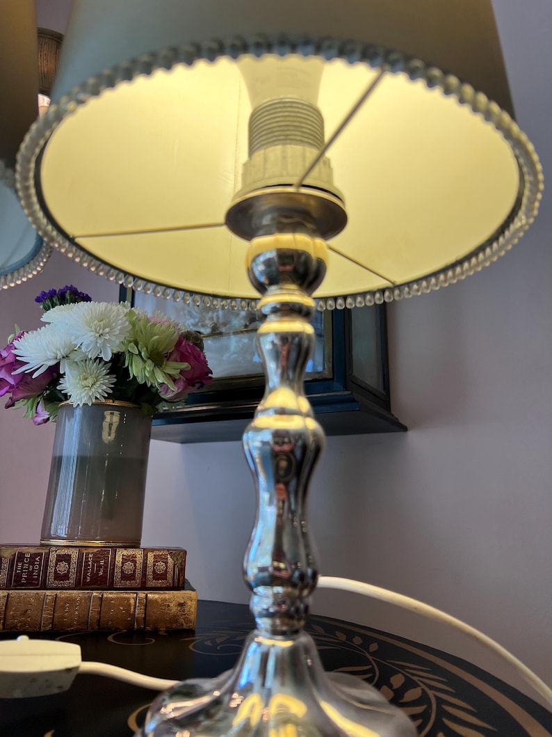 K&ouml;nnte beinhalten: Eine silberne Tischlampe mit beigem Lampenschirm und Perlenbesatz. Die Lampe steht auf einem Tisch mit einer Blumenvase und einem Stapel B&uuml;cher. Die Lampe ist eingeschaltet und wirft ein warmes Licht.