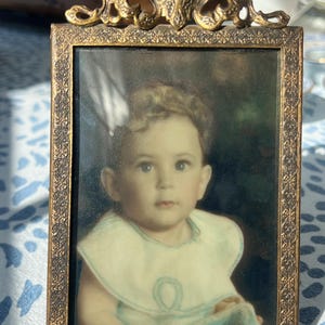 May include: A vintage gold-toned metal picture frame with an ornate design. The frame holds a faded photograph of a young child wearing a white bib.