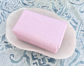 May include: A pink bar of soap with white glitter in a white ceramic dish. The soap is labeled "Bergamot & Spring Blossom" with a floral scent.