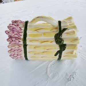 May include: A ceramic asparagus dish with a green ribbon and a handle. The asparagus is painted in shades of yellow, green, and purple.