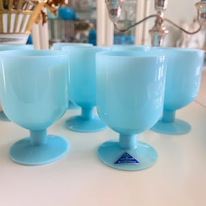 May include: A set of six light blue stemmed glasses. The glasses have a rounded bowl shape and a short stem. The glasses are made of a translucent material and are arranged on a white surface. A small blue and white label is visible on the base of one glass.