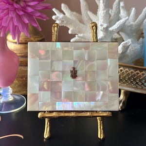 May include: A rectangular, mother-of-pearl mosaic box with a small emblem, displayed on a gold-colored easel. The box has gold-colored handles. The background includes a pink flower, a coral-like sculpture, and a pink vase.