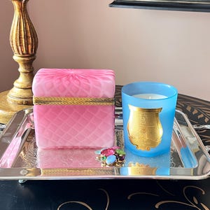 May include: A rectangular silver-toned tray holding a pink glass box with a diamond-patterned lid and gold-toned trim, a light blue candle in a glass jar with a gold-toned label, and a small cluster of multicolored glass decorative stones.