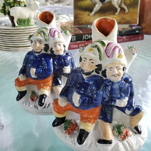 May include: Two ceramic figurines of men wearing blue jackets and red pants. They are sitting on a white base with green and red accents. The figurines are holding mugs and have a whimsical expression on their faces.
