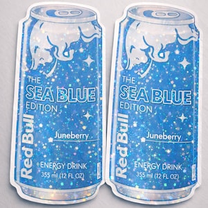 May include: Two stickers of Red Bull's Sea Blue Edition energy drink cans. The cans are blue with a glittery effect and feature the Red Bull logo, the flavor "Juneberry", and the text "ENERGY DRINK 355 ml (12 FL OZ)".