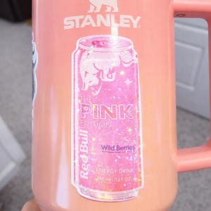 May include: A pink Stanley mug with a handle and a silver rim. A sticker of a pink Red Bull can is on the front, with the text "THE PINK EDITION" and "Wild Berries". The mug holds 12 fl oz.