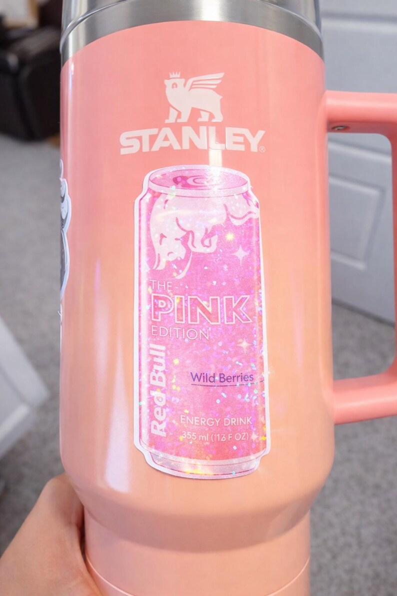May include: A pink Stanley mug with a handle and a silver rim. A sticker of a pink Red Bull can is on the front, with the text "THE PINK EDITION" and "Wild Berries". The mug holds 12 fl oz.