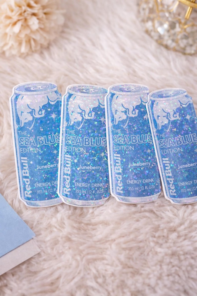 May include: Four stickers of Red Bull energy drink cans. The stickers are blue with glitter and feature the text "The Sea Blue Edition" and "Juneberry". Each can is 355 ml (12 fl oz).