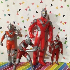 May include: Four Ultraman action figures in various poses, primarily red and silver, stand on a colorful striped surface. The figures vary in size, with a coin in the foreground. The background features a pattern of colorful triangles.
