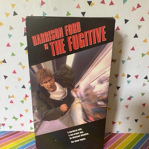 May include: A VHS tape of the movie "The Fugitive" starring Harrison Ford. The title is in red and white text on a black background. The image shows Ford running. The bottom of the tape has text that reads "A murdered wife. A one-armed man. An obsessed detective. The chase begins."