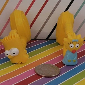 May include: Two yellow plastic Simpsons character figurines. The first is Bart Simpson with a blue shirt and the second is Lisa Simpson with a blue dress. The figurines are on a colorful striped background.