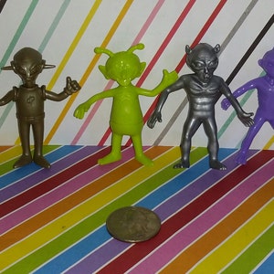 May include: Four plastic alien figurines in different colors: silver, green, purple, and brown. The figurines are standing on a striped background.