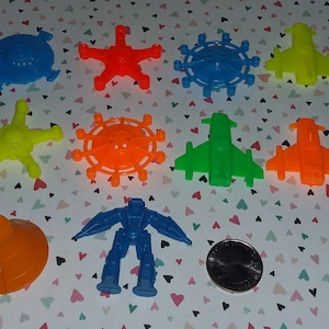 May include: A collection of ten colorful plastic toys, including a robot, a spaceship, a cone, and several star-shaped toys. The toys are arranged on a white background with a pink heart pattern.