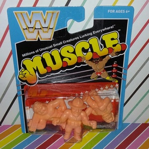 May include: A package of three small, orange, plastic wrestling figures. The figures are in a clear plastic blister pack. The package is labeled "WWE Muscle" and features a wrestler in a red and yellow outfit in a wrestling ring. The package is for ages 6 and up.