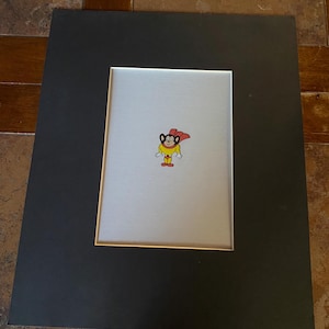 May include: Framed artwork featuring a cartoon character, Mighty Mouse, in a yellow outfit with a red cape. The artwork is set against a silver background, framed by a black mat and a gold inner border. The frame is black.