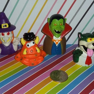 May include: Four Halloween finger puppets: a witch with a purple dress and a black hat, a green monster with orange skin and horns, a vampire with a brown cape and a blue shirt, and a werewolf with a green shirt and a red tie.