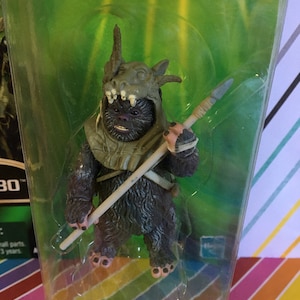 May include: A brown and grey action figure of a creature with a spear. The figure is wearing a green and brown helmet with horns. The figure is in a plastic package with a green background.