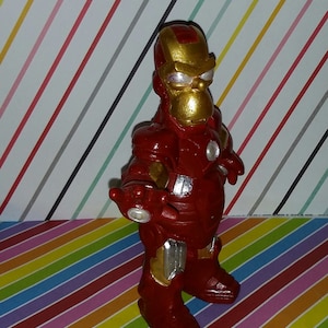 Op de afbeelding: Een handgemaakt beeldje van Iron Man, met een rood en goud kleurenschema. De figuur heeft een cartoonachtig uiterlijk, met een rond hoofd en een volumineus pak. De achtergrond is een kleurrijk strepenpatroon.