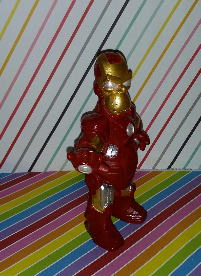 Op de afbeelding: Een handgemaakt beeldje van Iron Man, met een rood en goud kleurenschema. De figuur heeft een cartoonachtig uiterlijk, met een rond hoofd en een volumineus pak. De achtergrond is een kleurrijk strepenpatroon.