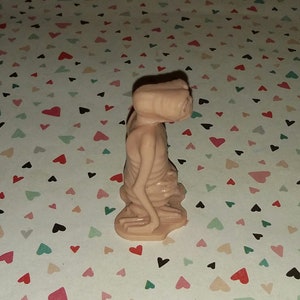 May include: A small, pink plastic figurine of an alien with large eyes and a long neck. The figurine is standing on a white background with a pattern of pink hearts.