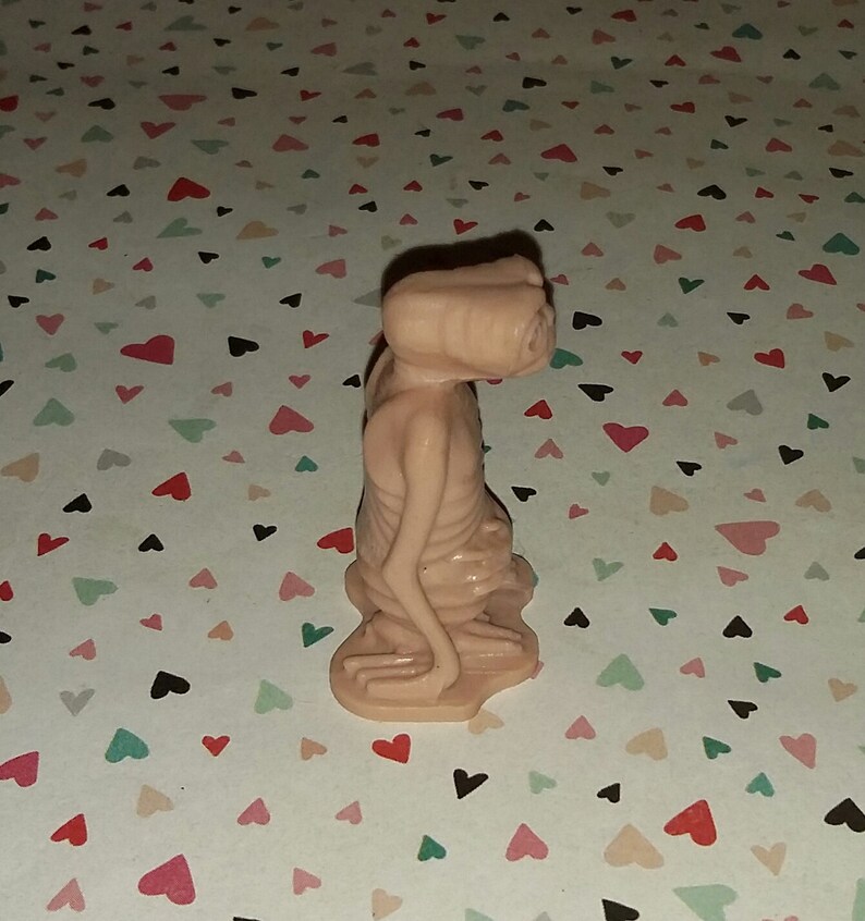 May include: A small, pink plastic figurine of an alien with large eyes and a long neck. The figurine is standing on a white background with a pattern of pink hearts.
