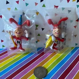 May include: Two red plastic Noid character figurines sealed in clear plastic bags. Each figure features a white letter N on the chest, white gloves, and a smiling face. One holds a yellow tool, the other a yellow staff. A US quarter is placed below.