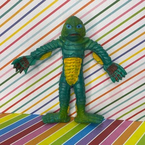 May include: A green and yellow monster figure with blue eyes and webbed hands and feet. The figure has a textured, scaly appearance and is posed with arms outstretched. The figure is a vintage toy.