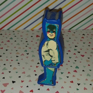 May include: Blue plastic toy figure of Batman. The figure is a cutout of Batman from a comic book. The figure is standing on a white background with a pattern of small hearts.