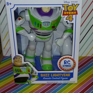 K&ouml;nnte beinhalten: Eine wei&szlig;e Box mit einer gr&uuml;n-wei&szlig;-violetten Buzz Lightyear Actionfigur. Auf der Box steht "Toy Story 4" und "RC 27MHZ". Die Figur h&auml;lt eine Laserpistole und tr&auml;gt einen gr&uuml;n-wei&szlig;en Space Ranger Anzug.