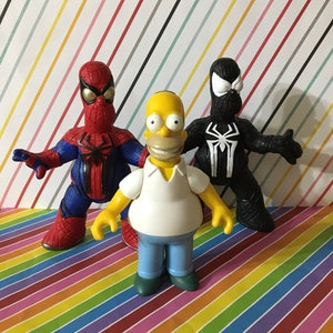 Peut inclure: Une figurine de dessin anim&eacute; jaune avec une chemise blanche et un jean bleu se tient entre une figurine de Spiderman rouge et bleue et une figurine de Spiderman noire. Les figurines sont toutes des jouets en plastique.