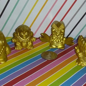 May include: Four gold-colored plastic toy monsters. The monsters are about 2 inches tall and have different designs. The monsters are on a colorful striped background.
