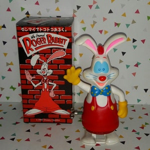 May include: A wind-up toy of Roger Rabbit from the film "Who Framed Roger Rabbit?" The toy is white with red overalls, a blue bow tie with white polka dots, and yellow gloves. The toy is standing on a white surface with a red brick background. The text "Who Framed Roger Rabbit?" is on the box behind the toy.