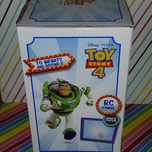 K&ouml;nnte beinhalten: Verpackung der Buzz Lightyear Fernbedienungsfigur aus Toy Story 4. Die Schachtel zeigt ein Cartoon-Bild von Buzz Lightyear in seinem Weltraum-Ranger-Anzug, der mit ausgestreckten Armen rennt. Die Schachtel ist wei&szlig; mit blauen und roten Akzenten. Der Text auf der Schachtel lautet "To Infinity and Beyond!" und "RC 27MHz Remote Control".