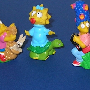 May include: A collection of five Simpsons character figurines. Homer wears a blue hat and green shirt. Lisa rides a snail. Maggie sits on a turtle. Marge holds a camera. Bart carries a skateboard and backpack. The figurines are brightly colored.