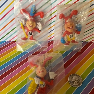 May include: Three small plastic figurines of a red cartoon character with a large head and long ears. The character is wearing a red and white suit with a large 'N' on the chest. Each figurine is packaged in a clear plastic bag. The character is shown in three different poses: holding a wand, wearing boxing gloves, and holding a drill.