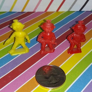 May include: Three small plastic astronaut figurines, one yellow, two red, standing on a striped background with a US quarter for scale.