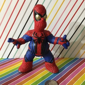 Peut inclure: Une figurine d'action Spider-Man rouge et bleue avec un visage jaune et un motif d'araign&eacute;e noir sur la poitrine. La figurine est debout sur un fond ray&eacute; color&eacute;.