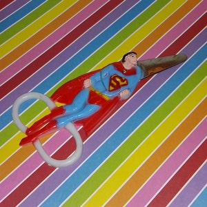 May include: A pair of white plastic scissors with a Superman figure attached to the blades. The figure is wearing a red cape and blue suit with the Superman logo on the chest. The scissors are on a colorful striped background.
