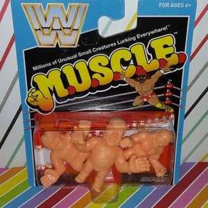 May include: A package of three small, orange, plastic wrestling figures. The figures are in a clear plastic blister pack on a red and white background. The package has a blue header with the WWF logo and the words "Millions of Unusual Small Creatures Lurking Everywhere!" in white text. The package also has the words "MUSCLE" in yellow text on a black background. The package is labeled "FOR AGES 6+".