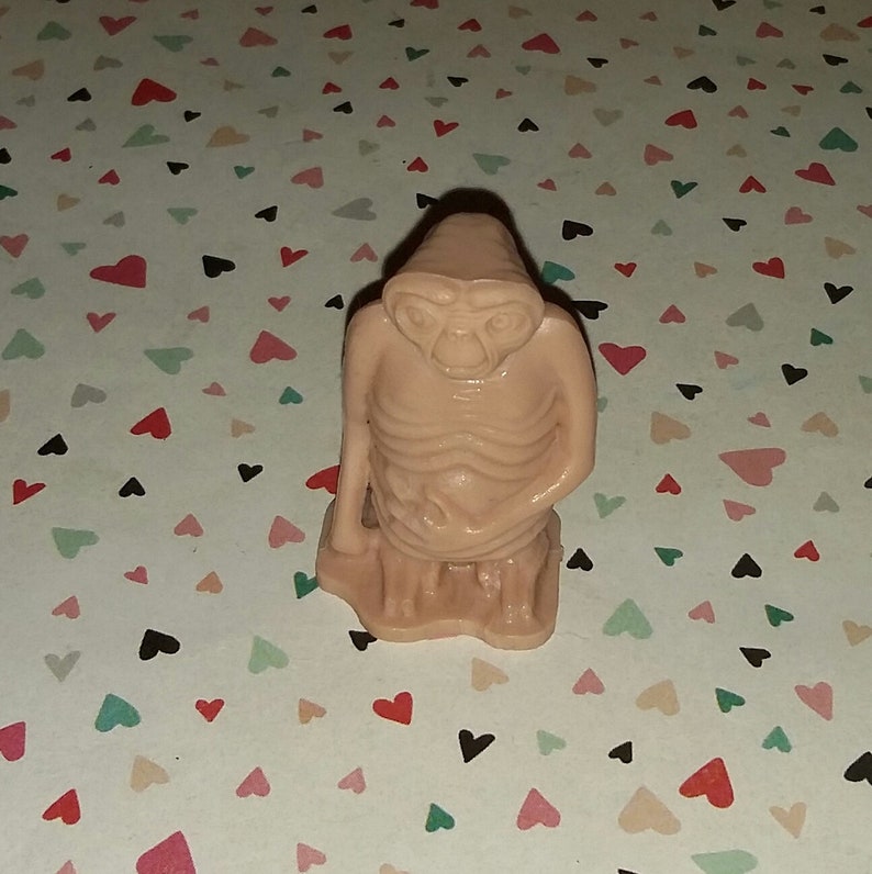 May include: A small, brown, plastic figurine of an alien with large eyes and a thin body. The figurine is standing on a base.