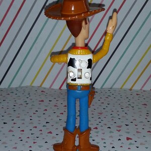 May include: A plastic toy figure of Woody from Toy Story. Woody is wearing his signature brown cowboy hat, a yellow vest with a black and white cow pattern, blue jeans, and brown boots. He is waving his right arm and has a pull string on his back.