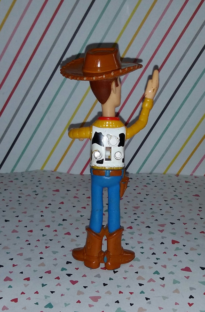 May include: A plastic toy figure of Woody from Toy Story. Woody is wearing his signature brown cowboy hat, a yellow vest with a black and white cow pattern, blue jeans, and brown boots. He is waving his right arm and has a pull string on his back.